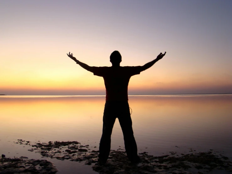 a man standing on top of a beach at sunset, arms spread wide, standing next to water, pray, standing straight