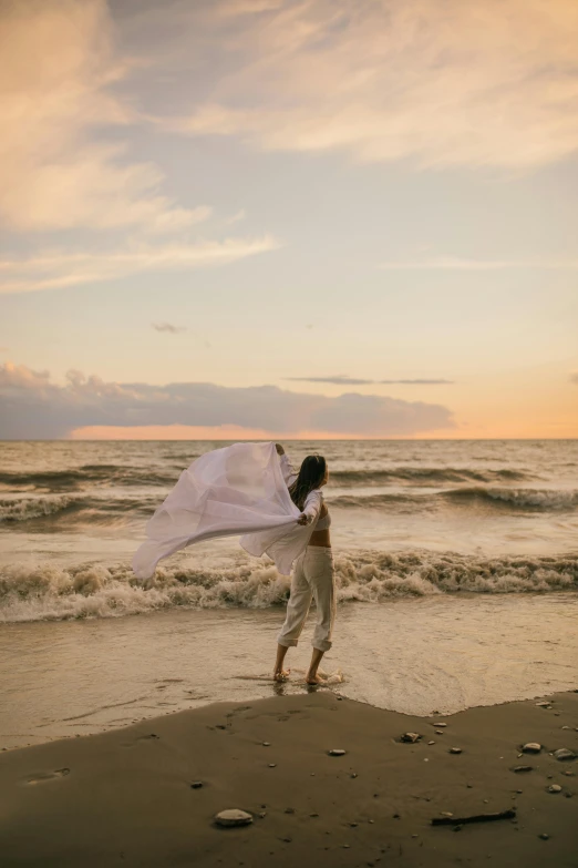 a woman standing on top of a beach next to the ocean, white cloth in wind shining, sunset at the beach, twirling, kailee mandel