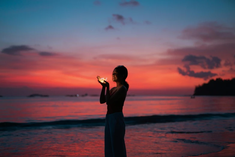 a woman standing on a beach holding a seashell, by Julia Pishtar, pexels contest winner, candle lighting, redpink sunset, standing on a beach in boracay, doing a prayer