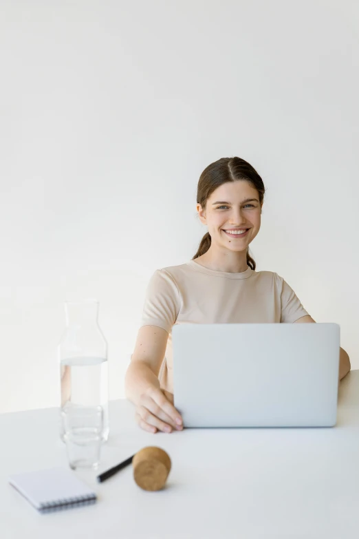 a woman sitting at a table with a laptop, plain background, smiling slightly, girl with brown hair, hydration