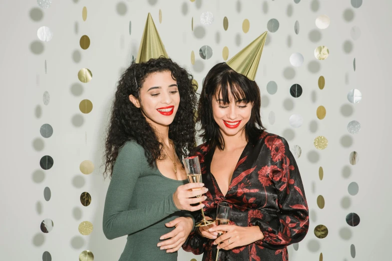 a couple of women standing next to each other, trending on pexels, wearing a party hat, backdrop, an olive skinned, new years eve