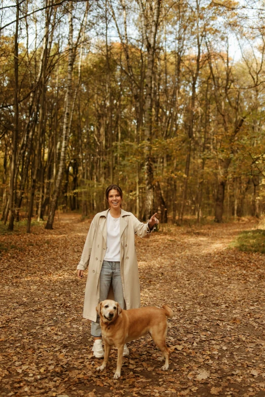 a woman standing in the woods with a dog, wearing a long beige trench coat, in louisiana, hermes, woodland location