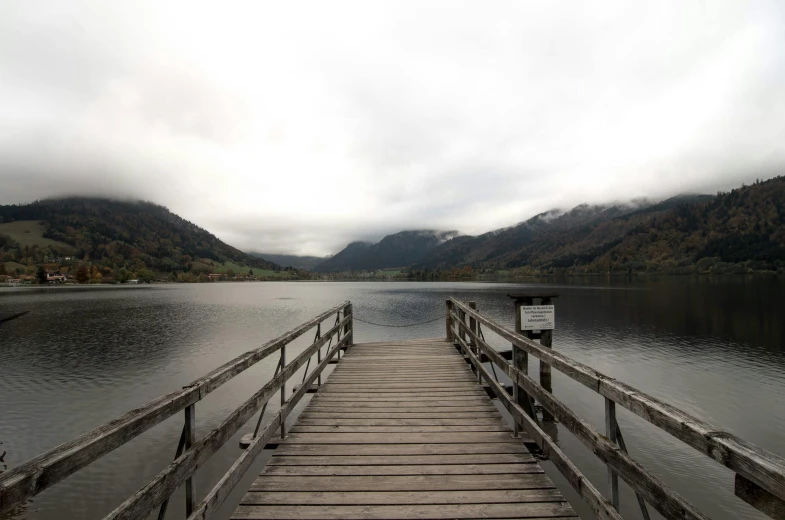 a dock on a lake with mountains in the background, by Otto Meyer-Amden, unsplash, overcast gray skies, wooden bridge, haunted, festivals