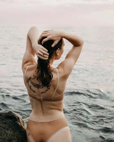a woman in a bikini sitting on a rock by the ocean, a tattoo, by Grace Polit, trending on pexels, renaissance, rippling muscles, arched back, non binary model, with arms up