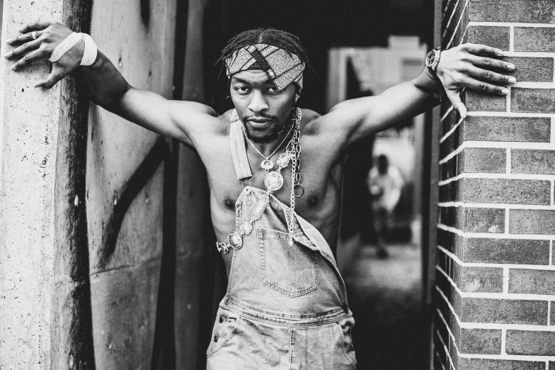 a black and white photo of a man leaning against a brick wall, an album cover, inspired by Ras Akyem, trending on pexels, renaissance, wearing a bandana and chain, artist wearing torn overalls, short dreadlocks with beads, sassy pose