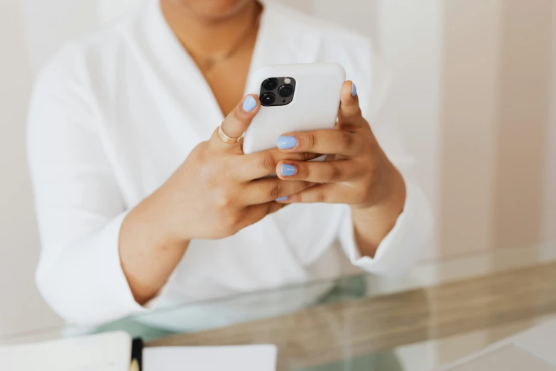 a close up of a person holding a cell phone, by Julia Pishtar, trending on pexels, wearing white cloths, on a desk, with brown skin, manicured