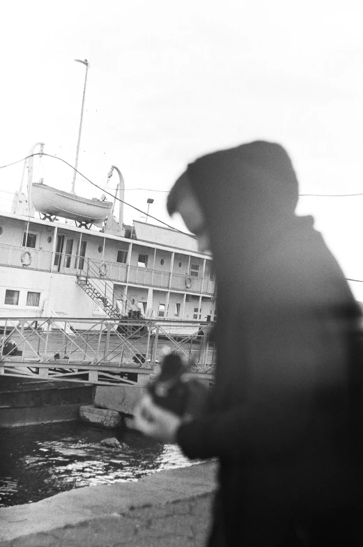 a man standing next to a body of water with a boat in the background, a black and white photo, by Eric Peterson, hooded figure, on the deck of a ship, ( ( abstract ) ), musician