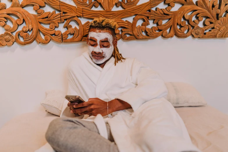 a man in a white robe sitting on a bed, a photo, trending on pexels, happening, tribal mask, skincare, avatar image, with facial tattoo