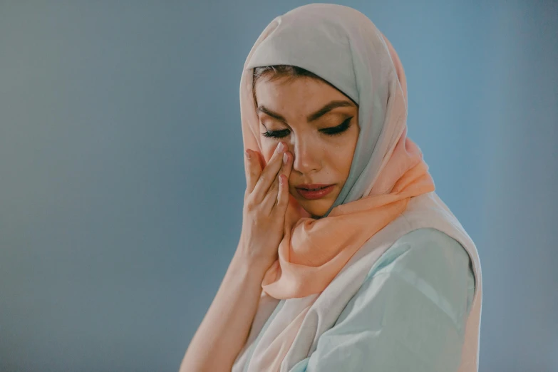 a woman in a hijab covers her face with her hands, a colorized photo, trending on pexels, hurufiyya, pastel blues and pinks, looking sad, background image, in shades of peach