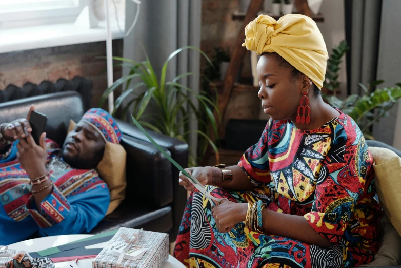 a woman sitting on a couch next to a man on a cell phone, by Alice Mason, pexels contest winner, afrofuturism, cloth head wrap, still from a live action movie, colourful clothes, at home