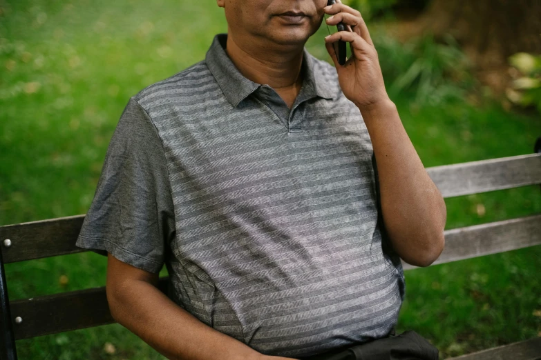 a man sitting on a bench talking on a cell phone, gray shirt, nivanh chanthara, wearing polo shirt, an oldman