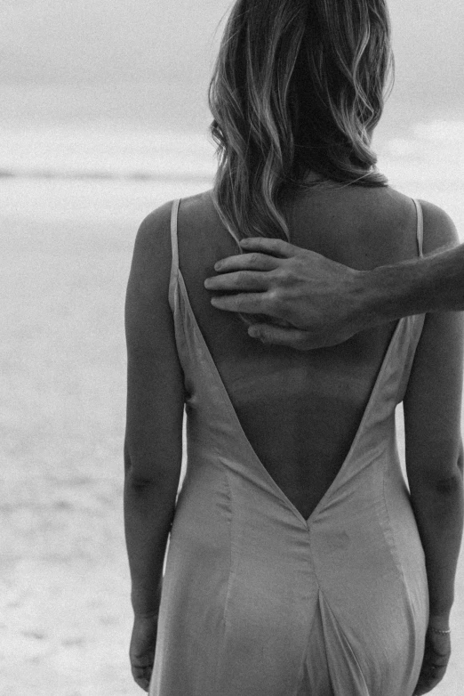 a woman standing on top of a beach next to the ocean, a black and white photo, tumblr, man grabbing a womans waist, open back dress, zoomed in, aesthetics