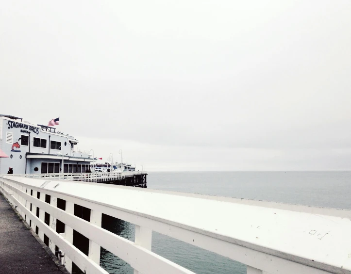 a boat sitting on top of a pier next to a body of water, by Carey Morris, unsplash, postminimalism, pure white hazy overcast sky, the city of santa barbara, on the deck of a ship, photographic print