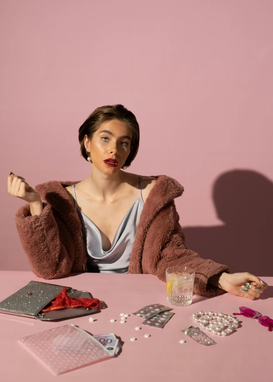 a woman sitting at a table with pills and pills, an album cover, trending on pexels, magic realism, lily collins, wearing a grey fur robe, wearing a light - pink suit, non binary model