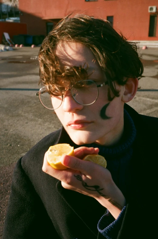 a close up of a person holding a piece of food, an album cover, unsplash, bauhaus, attractive androgynous humanoid, teen boy, circular sunglasses, moon behind him
