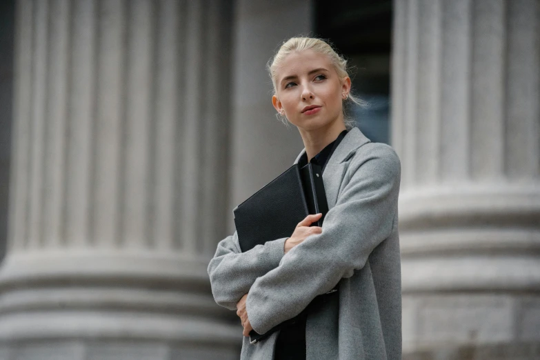 a woman standing in front of a building holding a book, lawyer clothing, grey jacket, young blonde woman, 2019 trending photo