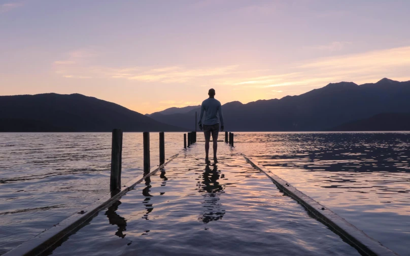 a man standing on a dock in the middle of a lake, by Sophie Pemberton, pexels contest winner, at purple sunset, new zealand, album art, lake kawaguchi