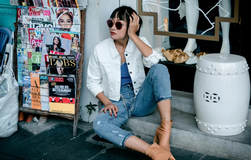 a woman sitting on the steps of a building, wearing a jeans jackets, milk bar magazine, circular sunglasses, set on singaporean aesthetic