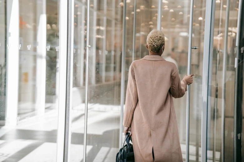 a woman standing in front of a glass door, light brown coat, busy, reuniting, glass ceilings