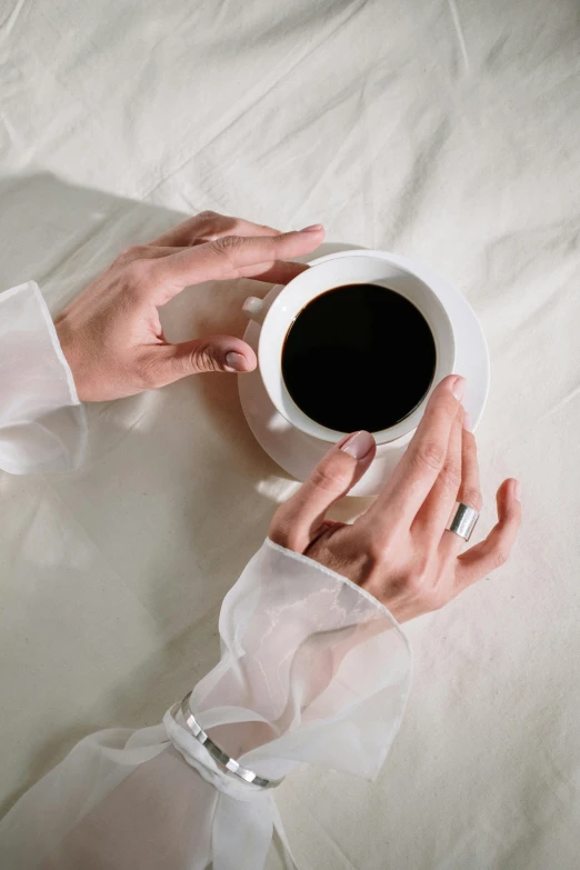 a close up of a person holding a cup of coffee, black nails, sitting on the edge of a bed, ring lit, on a white table