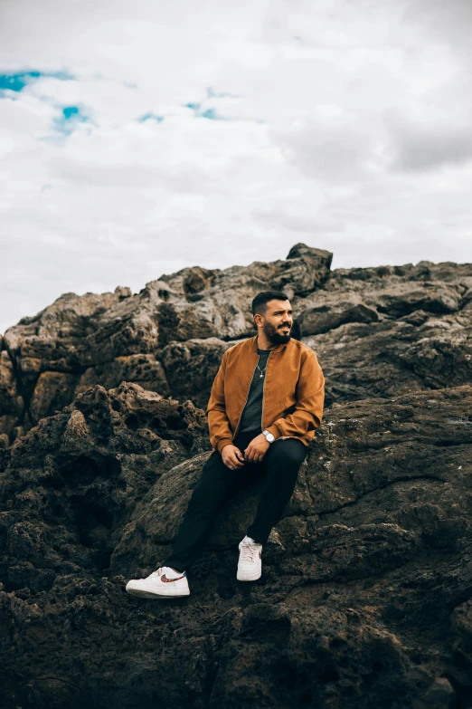 a man sitting on top of a pile of rocks, an album cover, trending on unsplash, hurufiyya, a portrait of rahul kohli, in australia, wearing a bomber jacket, ocean view