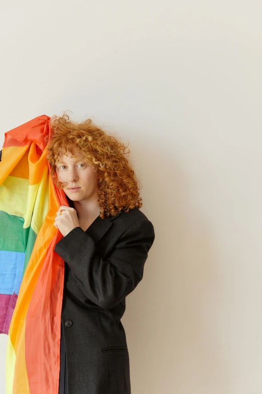 a woman standing in front of a white wall holding a rainbow blanket, an album cover, by Harriet Zeitlin, with curly red hair, holding a white flag, half male and half female, slide show