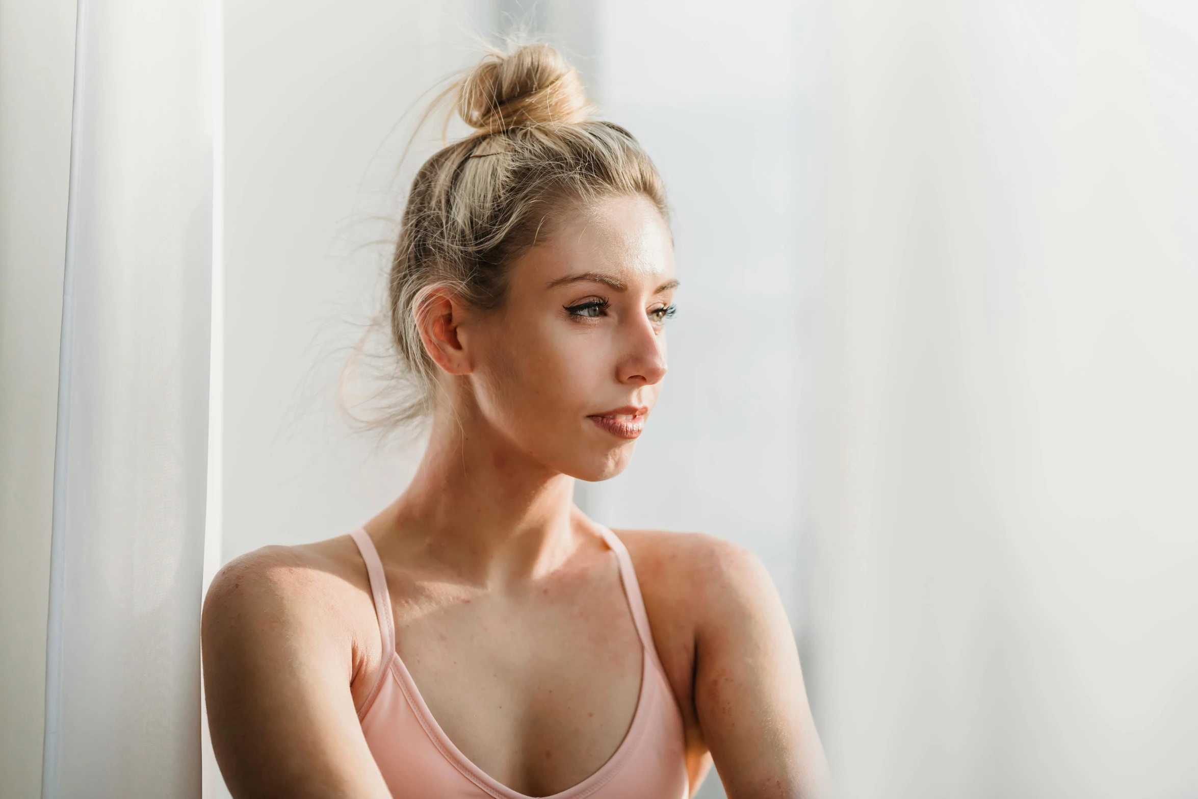 a woman in a pink top leaning against a wall, trending on pexels, tight bun, looking out window, blonde and attractive features, clean face and body skin