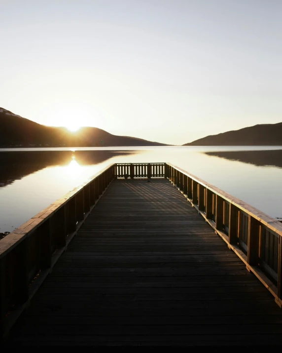 a dock in the middle of a lake at sunset, by Jessie Algie, unsplash, hurufiyya, sun rises between two mountains, walkway, fjords, deserted