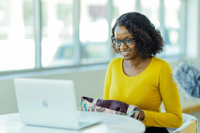 a woman sitting at a table with a laptop, by Ella Guru, pexels contest winner, with yellow cloths, ai researcher, student, holding a gold bag