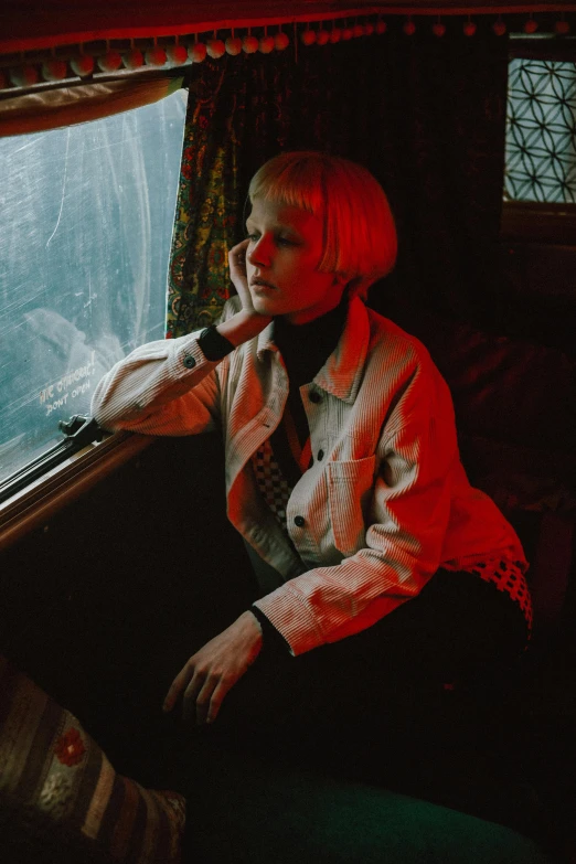 a woman sitting on a train looking out a window, an album cover, inspired by Elsa Bleda, short platinum hair tomboy, with red haze, lily frank, she wears a jacket