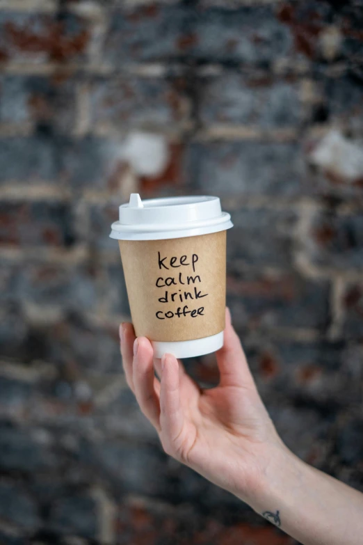a person holding a cup of coffee in front of a brick wall, text on paper, paper cup, thumbnail, calm