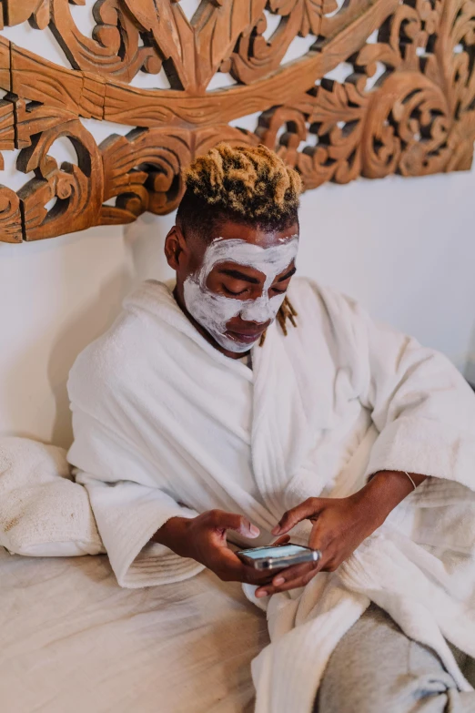 a man sitting on a bed using a cell phone, trending on pexels, afrofuturism, white face paint, bali, spa, hair styled in a bun