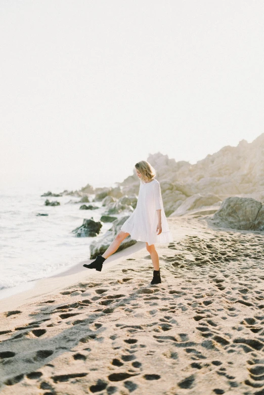 a woman in a white dress walking on a beach, unsplash, happening, white blouse and gothic boots, vsco film grain, a blond, celebrating