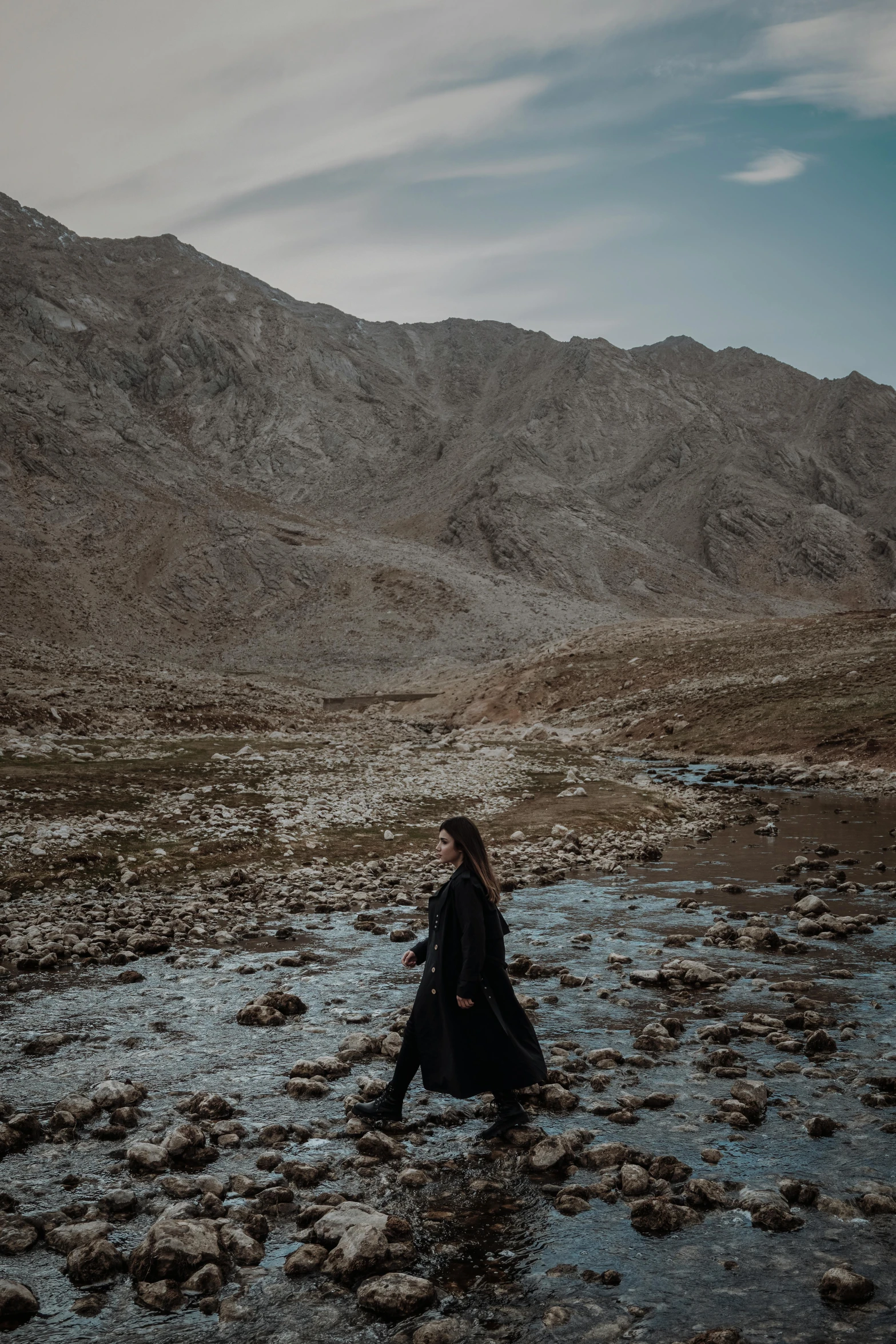 a person standing in a river with mountains in the background, an album cover, unsplash contest winner, les nabis, woman in black robes, kurdistan, standing on rocky ground, cinematic outfit photo