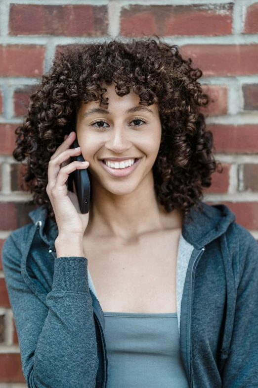 a woman standing in front of a brick wall talking on a cell phone, by Washington Allston, trending on pexels, renaissance, curly bangs, official product photo, smiling young woman, east african man with curly hair