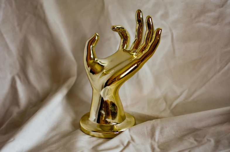 a golden hand statue sitting on top of a white sheet, a surrealist sculpture, inspired by Méret Oppenheim, unsplash, new sculpture, brass plated, high quality product photo, palm body, shiny golden
