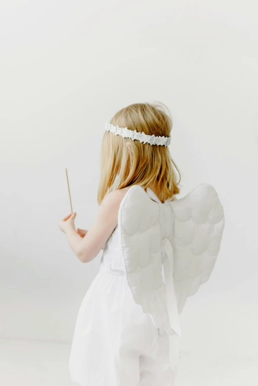 a little girl dressed as an angel holding a wand, light grey backdrop, back towards camera, full product shot, wearing a paper crown