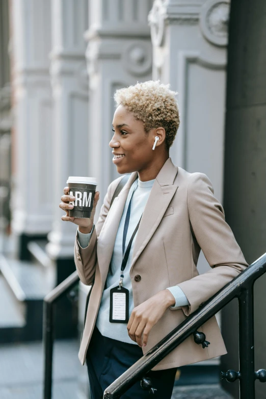 a woman standing on the steps of a building holding a cup of coffee, by Carey Morris, trending on unsplash, wearing a blazer, earbuds jewelry, short blonde afro, holding arms on holsters