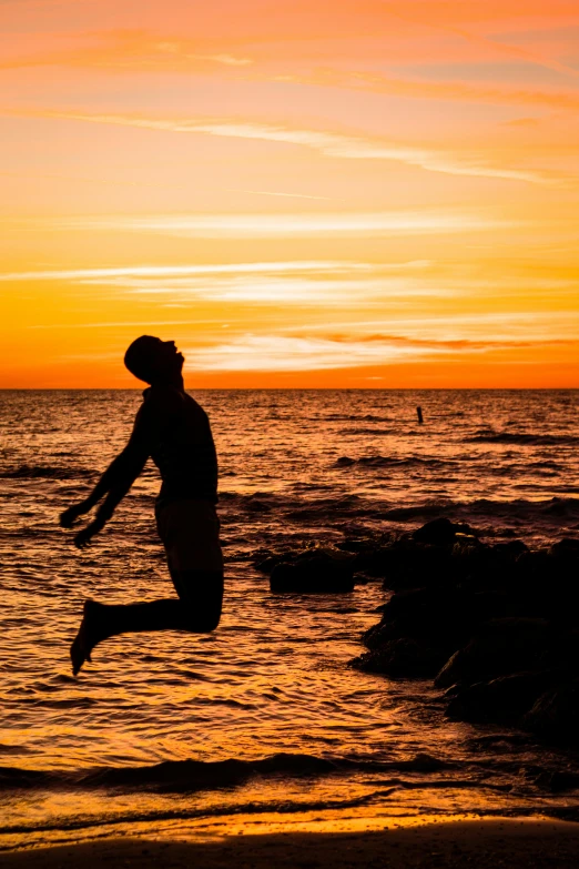 a person jumping in the air on a beach, during a sunset, emerging from the water, during sunset, manly