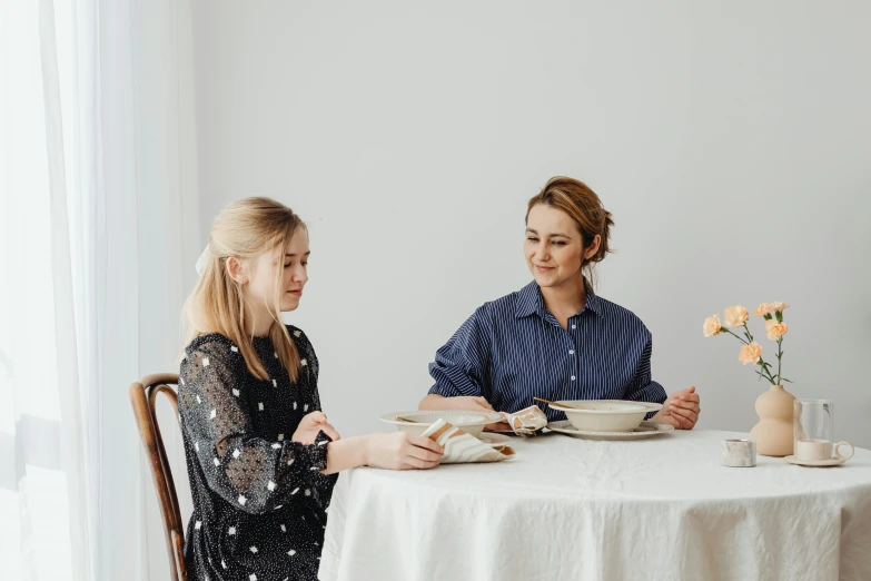 two women sitting at a table with plates of food, a portrait, by Alice Mason, pexels contest winner, private press, set against a white background, linen, two girls, background image