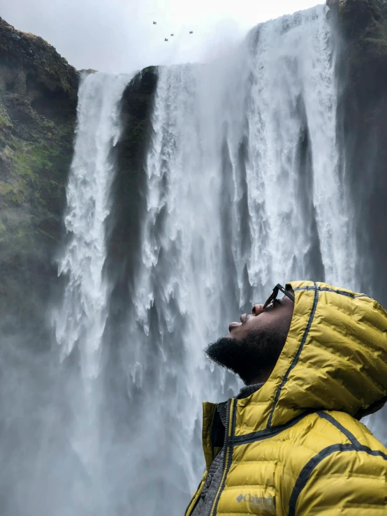 a man standing in front of a waterfall, inspired by national geographic, pexels contest winner, wearing a yellow hoodie, black man with afro hair, iceland, hood covers his eyes