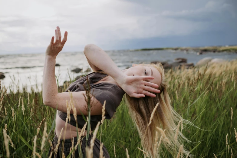 a woman standing in tall grass near a body of water, by Christen Dalsgaard, unsplash, magic realism, holding his hands up to his face, as though she is dancing, in an action pose, a girl with blonde hair