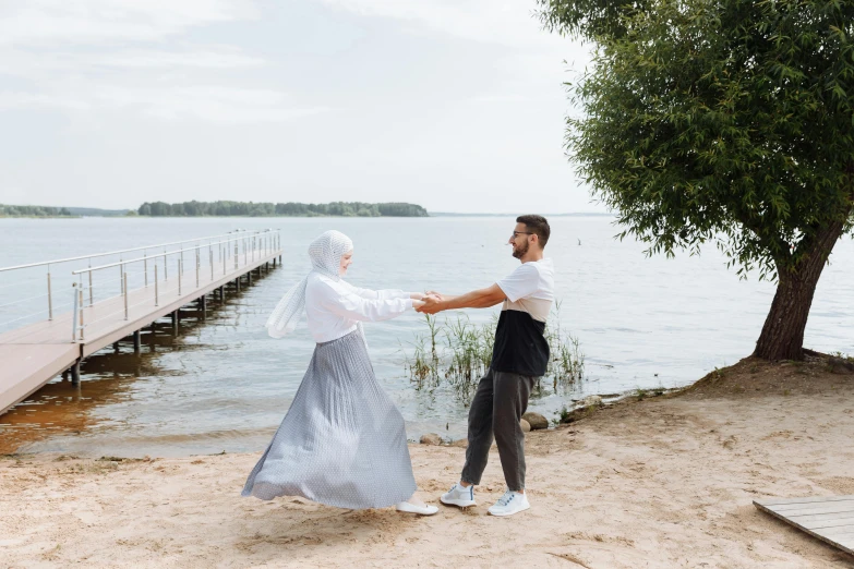 a man standing next to a woman on a beach, by Grytė Pintukaitė, pexels contest winner, hurufiyya, hijab, dancing elegantly over you, lakeside, white shirt and grey skirt