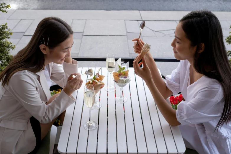 two women sitting at a table eating food, by Nina Hamnett, pexels contest winner, champagne commercial, white, exterior shot, reverse