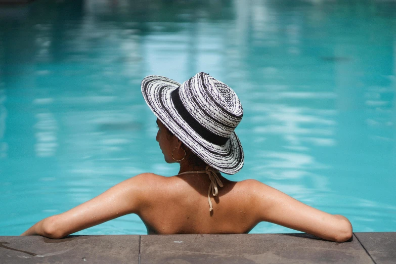 a woman sitting at the edge of a swimming pool, pexels contest winner, wearing wide sunhat, showing her shoulder from back, striped, profile image