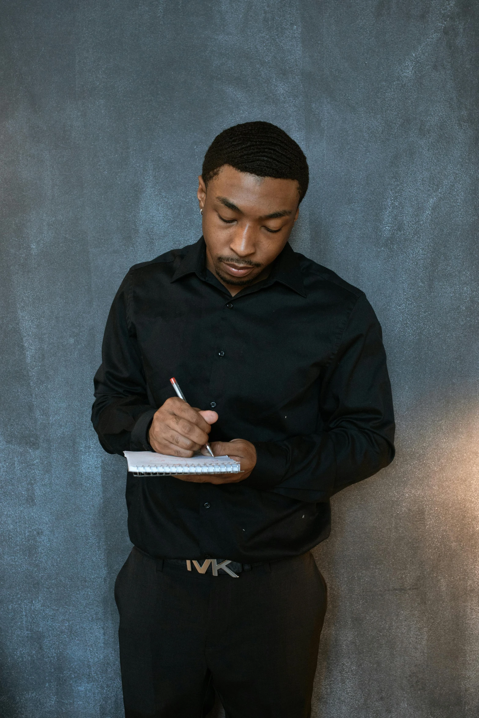 a man standing in front of a wall writing on a piece of paper, inspired by Kevin A. Short, mkbhd, wearing a black shirt, ☁🌪🌙👩🏾, holding a book