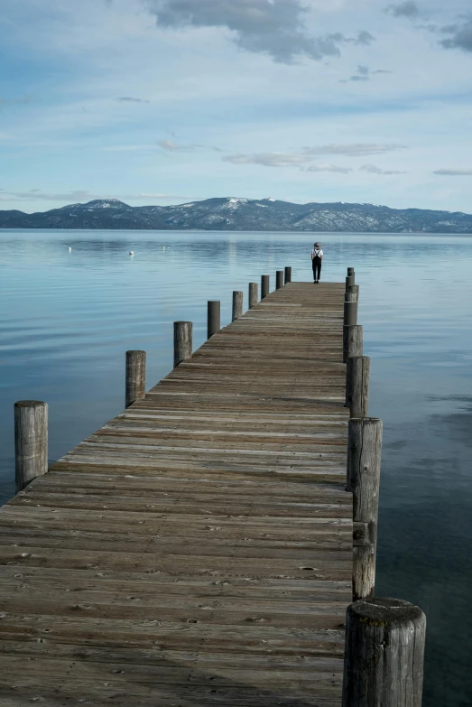 a wooden pier stretching out into the water, inspired by Frank Weston Benson, lone person in the distance, peaks, bay, heavenly