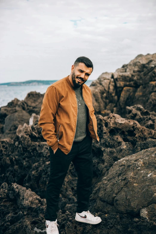 a man standing on rocks near the ocean, a picture, inspired by Harrington Mann, renaissance, brown leather jacket, stubble, drake, smart casual