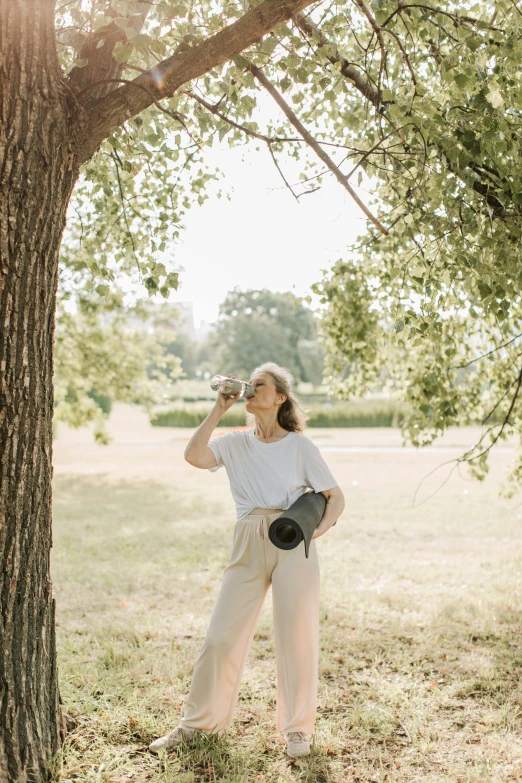 a woman standing under a tree talking on a cell phone, unsplash contest winner, happening, full body potrait holding bottle, yoga, white wine bottle, wearing pants and a t-shirt