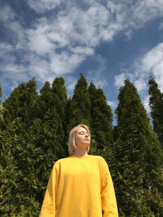 a woman standing in front of a row of trees, an album cover, by Attila Meszlenyi, wearing a modern yellow tshirt, wearing an oversized sweater, sky view, blonde swedish woman